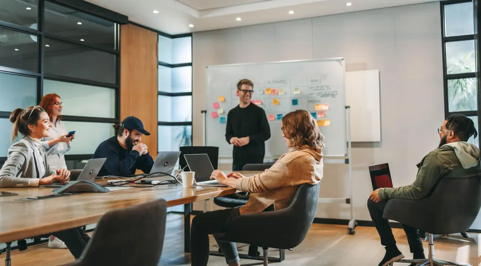 Team meeting in a bright office: a man stands at a whiteboard with colorful sticky notes while colleagues work on laptops.