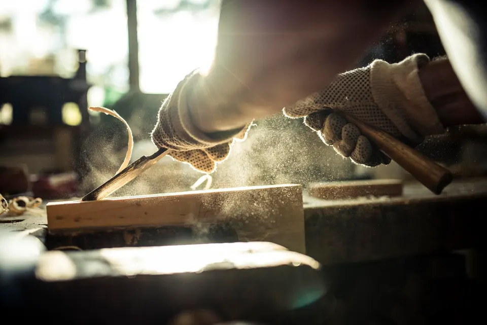 0x0 (32) Two gloved hands carve a wooden block with a chisel as sawdust rises in a sunlit workshop.